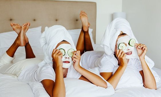 Dvije ženske osobe leže na krevetu u bjelim majcama, ručnicima na glavi a na licu piling za lice i kriške krastavca na očima